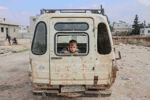 pexels-photo-6929741-6929741 A young boy peers from an abandoned van in the desolate streets of Idlib, Syria, amidst war devastation.