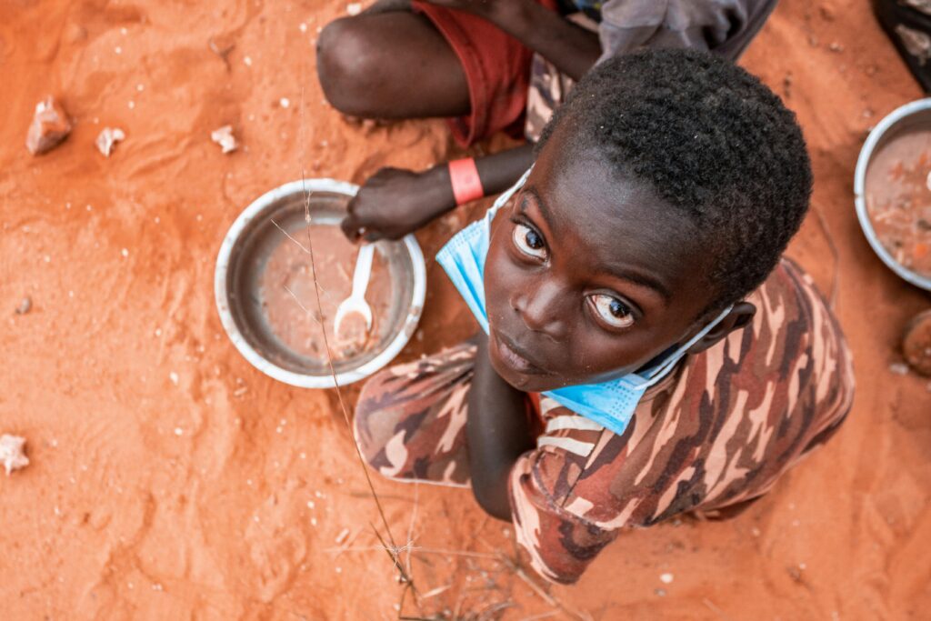 pexels-photo-10311711-10311711 Child on a sandy ground in Luanda with mask and empty bowl, highlighting pandemic challenges.